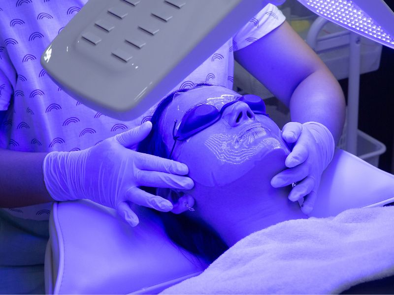 Client laying under Blue light led skin treatment in spa room with esthetician 